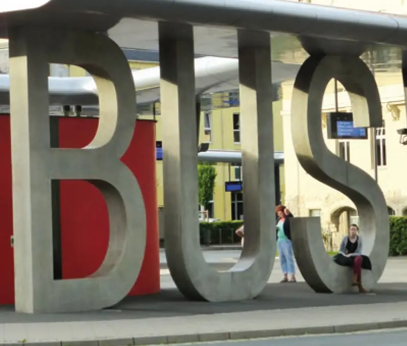 Bushaltestelle mit großen Betonbuchstaben, die das Wort 'BUS' bilden, und einer wartenden Person.