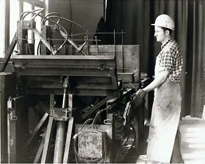 Ein Arbeiter in einem Helm steht neben einer großen Maschine in einer Fabrikumgebung.