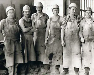 Sechs Handwerker in Arbeitskleidung und Schutzhelmen stehen nebeneinander in einer Werkstatt.