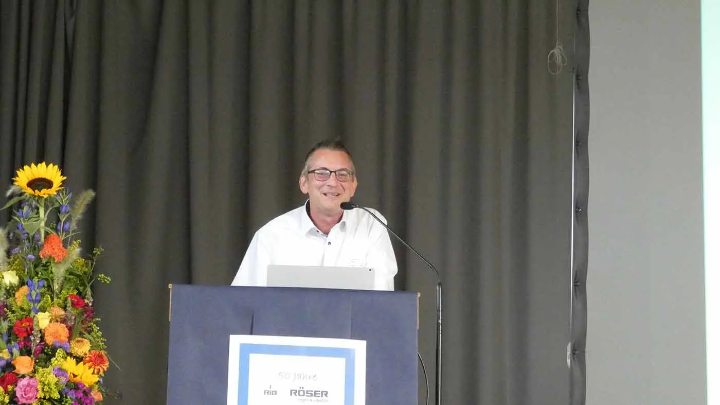 Mann spricht am Rednerpult während einer Veranstaltung mit einer Präsentation im Hintergrund.