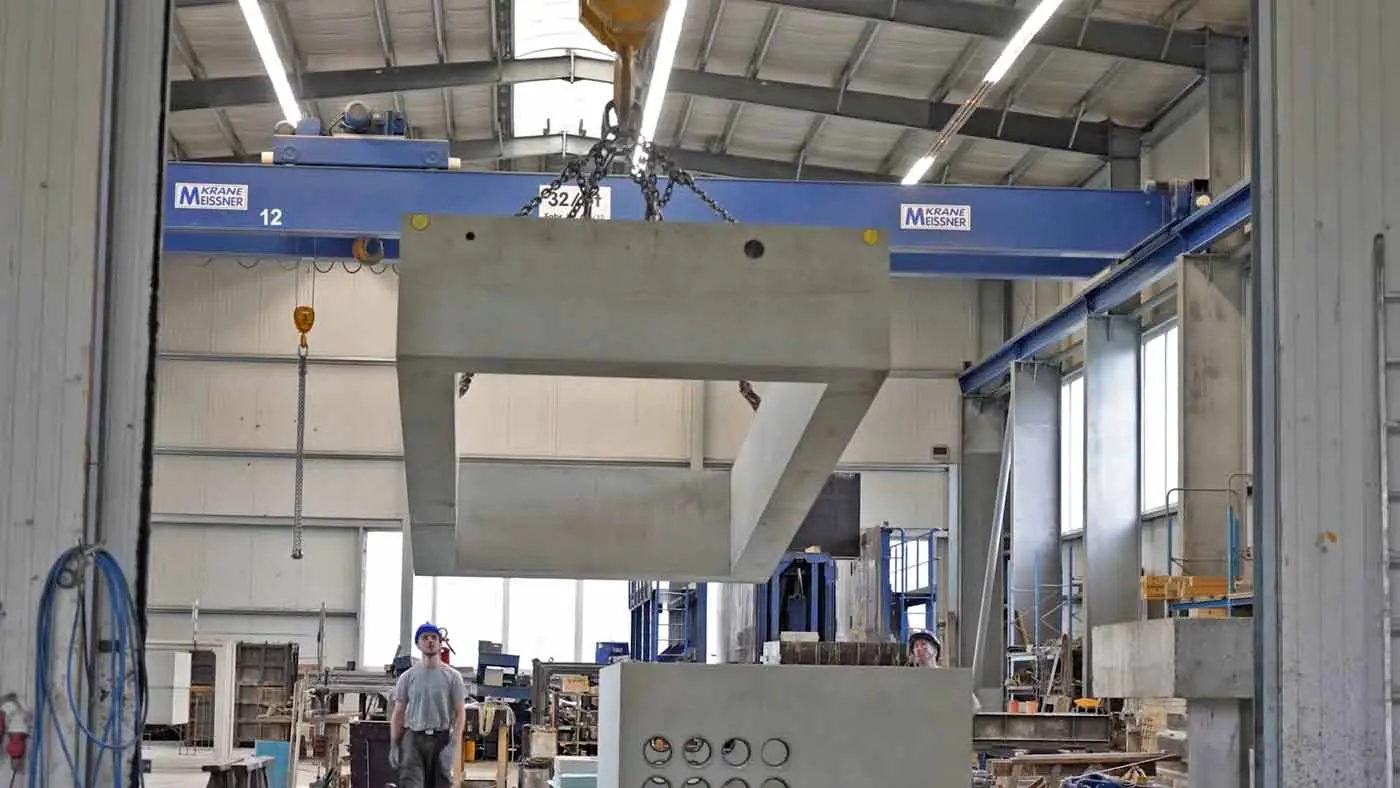 Ein Kran hebt einen großen Betonblock in einer Produktionshalle mit mehreren Arbeitern im Hintergrund.