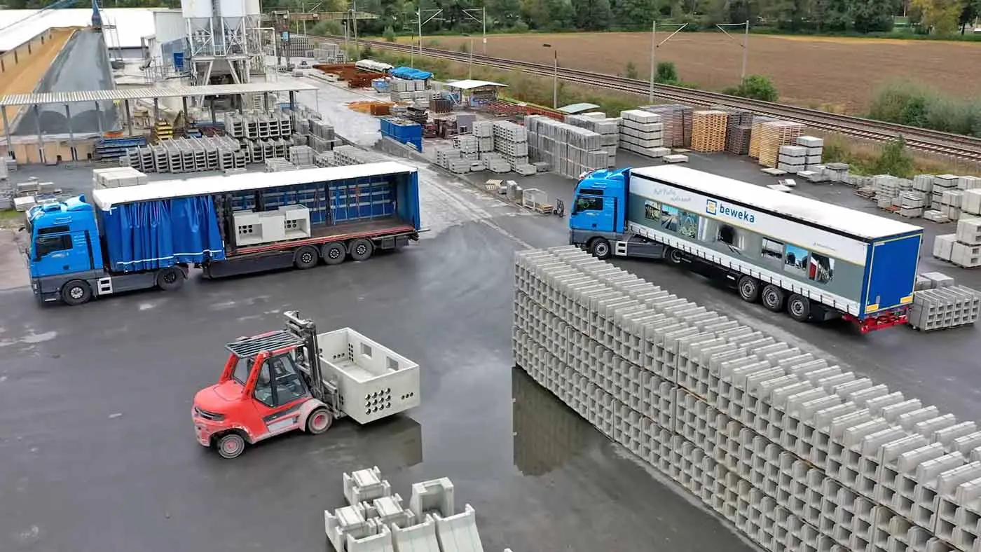 Zwei Lkw und ein Gabelstapler in einem Logistikbereich mit Betonsteinen.