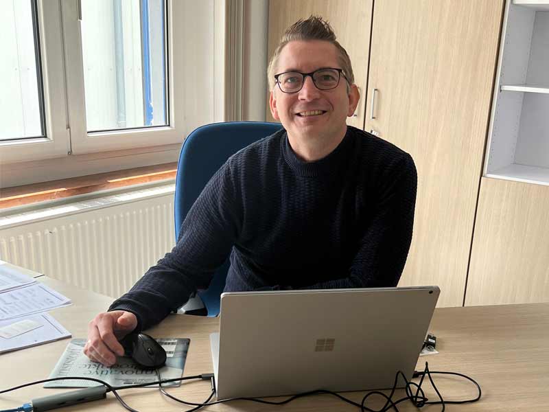 Frank Röser sitzt an einem Schreibtisch und arbeitet an einem Laptop mit einer Maus.