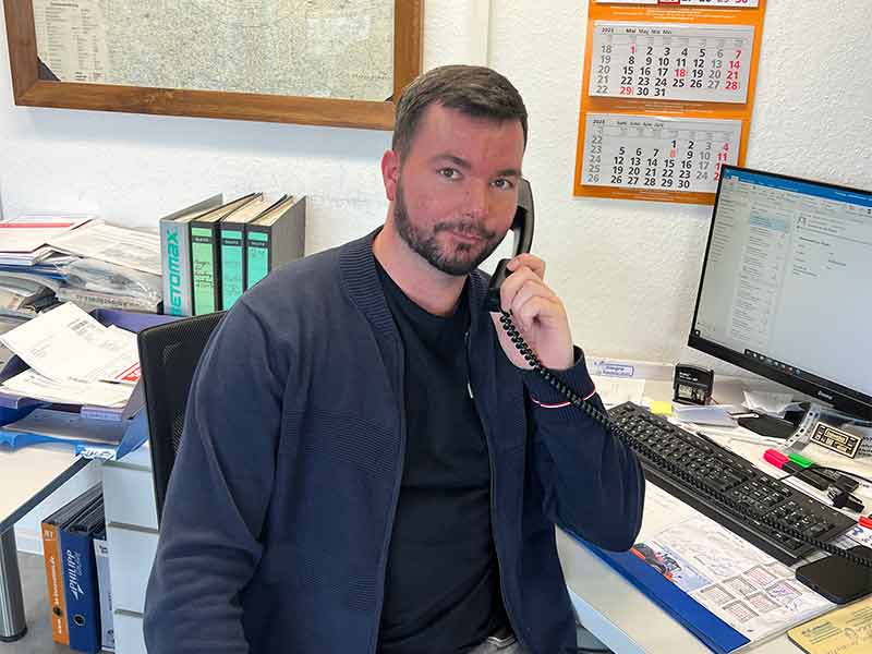 Mann sitzt am Schreibtisch und spricht am Telefon, umgeben von Bürounterlagen und einem Computer.