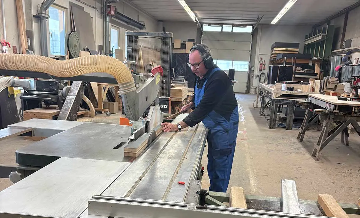 Ein Tischler arbeitet an einer Tischkreissäge in einer Werkstatt mit Holzmaterialien.